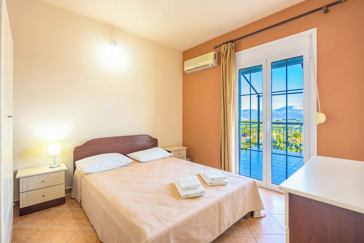 A bedroom with a bed, nightstand, lamp, air conditioning unit, and a balcony with a view.