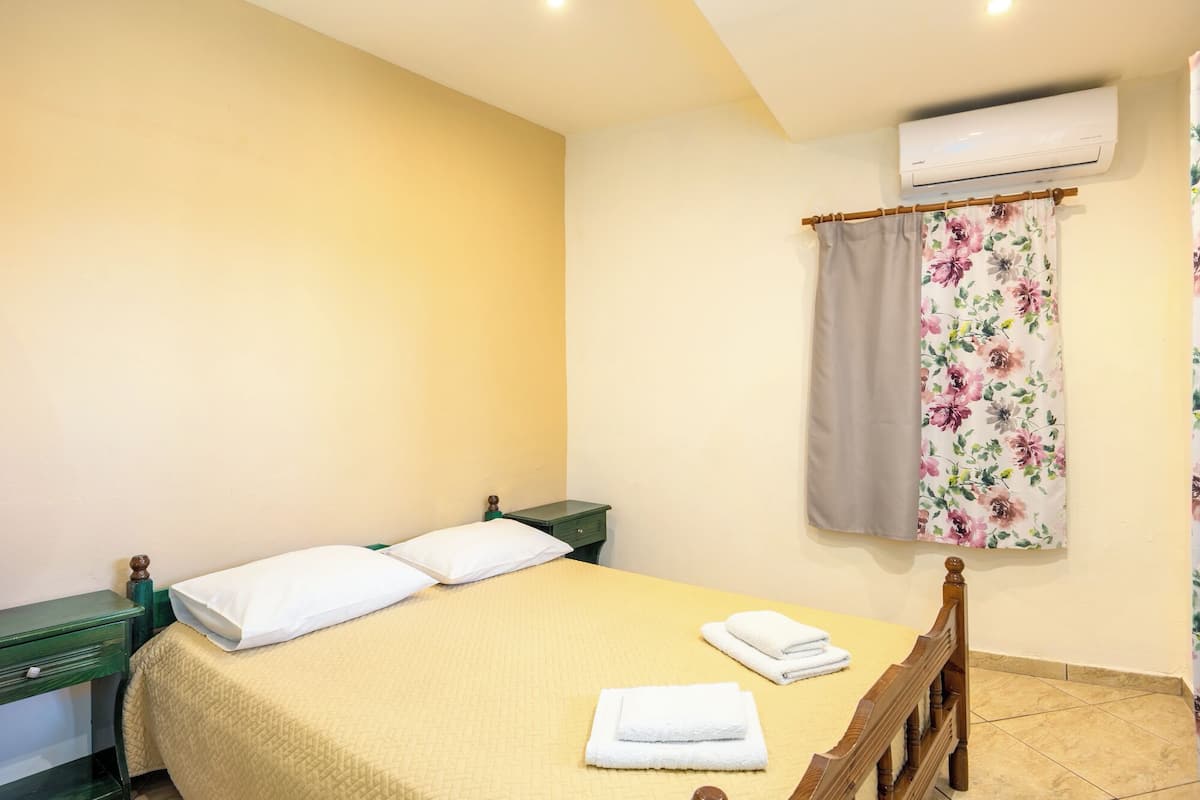 A bedroom with a bed, pillows, a bedside table, a chair, a curtain, and an air conditioning unit.