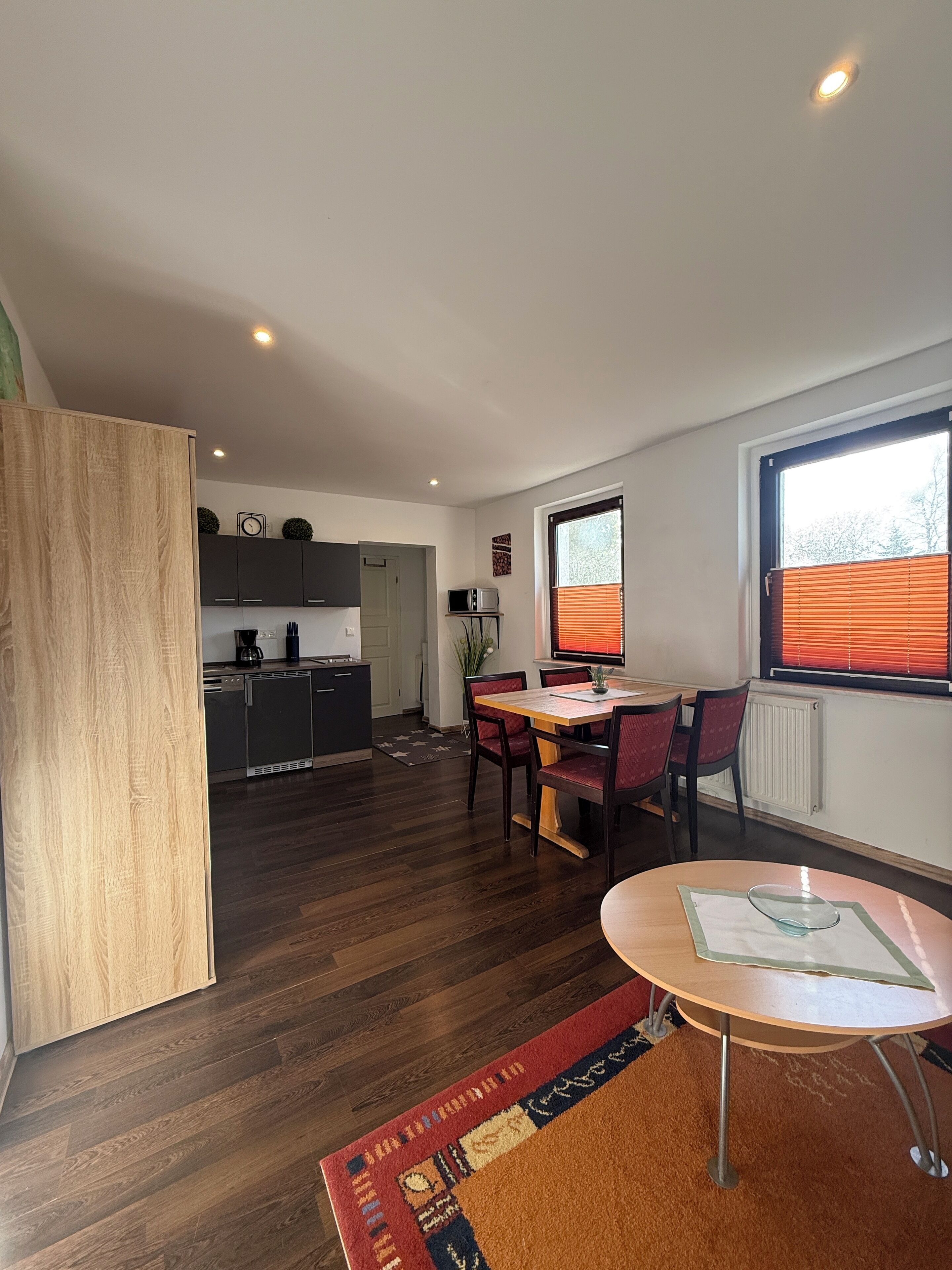 A modern kitchen with dark cabinets, a dining area with a wooden table and chairs, and a large window with blinds.