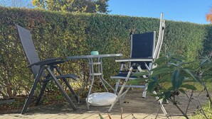 Un espace extérieur aménagé avec des chaises et une table en métal, entouré d’une haie et de plantes.
