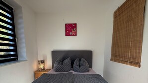 Une chambre à coucher comprenant un lit, des oreillers, une table de chevet, une lampe, un tableau décoratif et une fenêtre avec des stores.