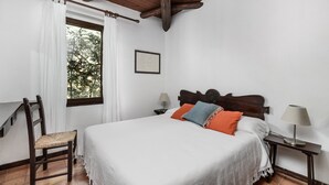 A bedroom with a wooden bed, white curtains, a wooden chair, a nightstand with a lamp, and a window with a view of trees. - Villa Romana – Beach House with Fenced Garden at Costa Rei (Costa Rei)