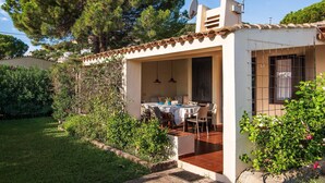 A small, single-story building with a patio area, a table with chairs, and a garden with various plants and trees. - Villa Romana – Beach House with Fenced Garden at Costa Rei (Costa Rei)