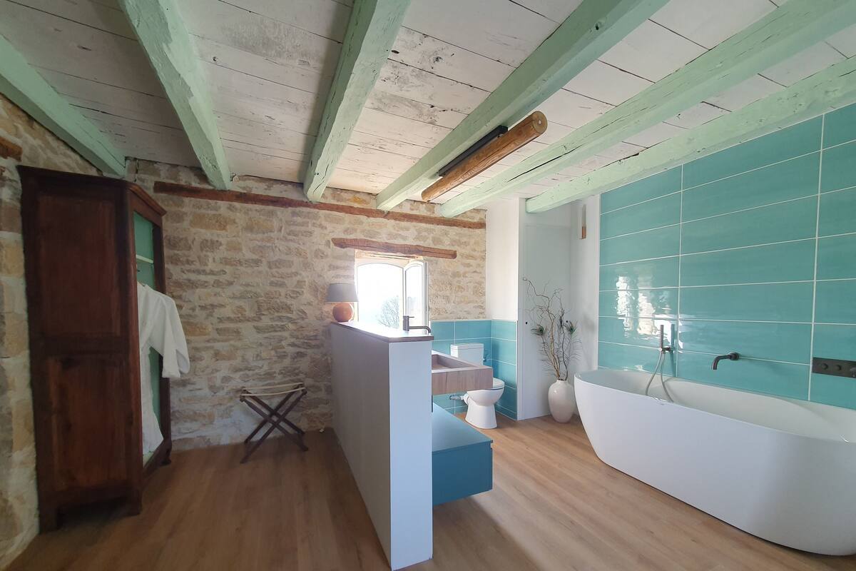 A bathroom with a wooden floor, a stone wall, a white sink, a toilet, and a bathtub with blue tiles.