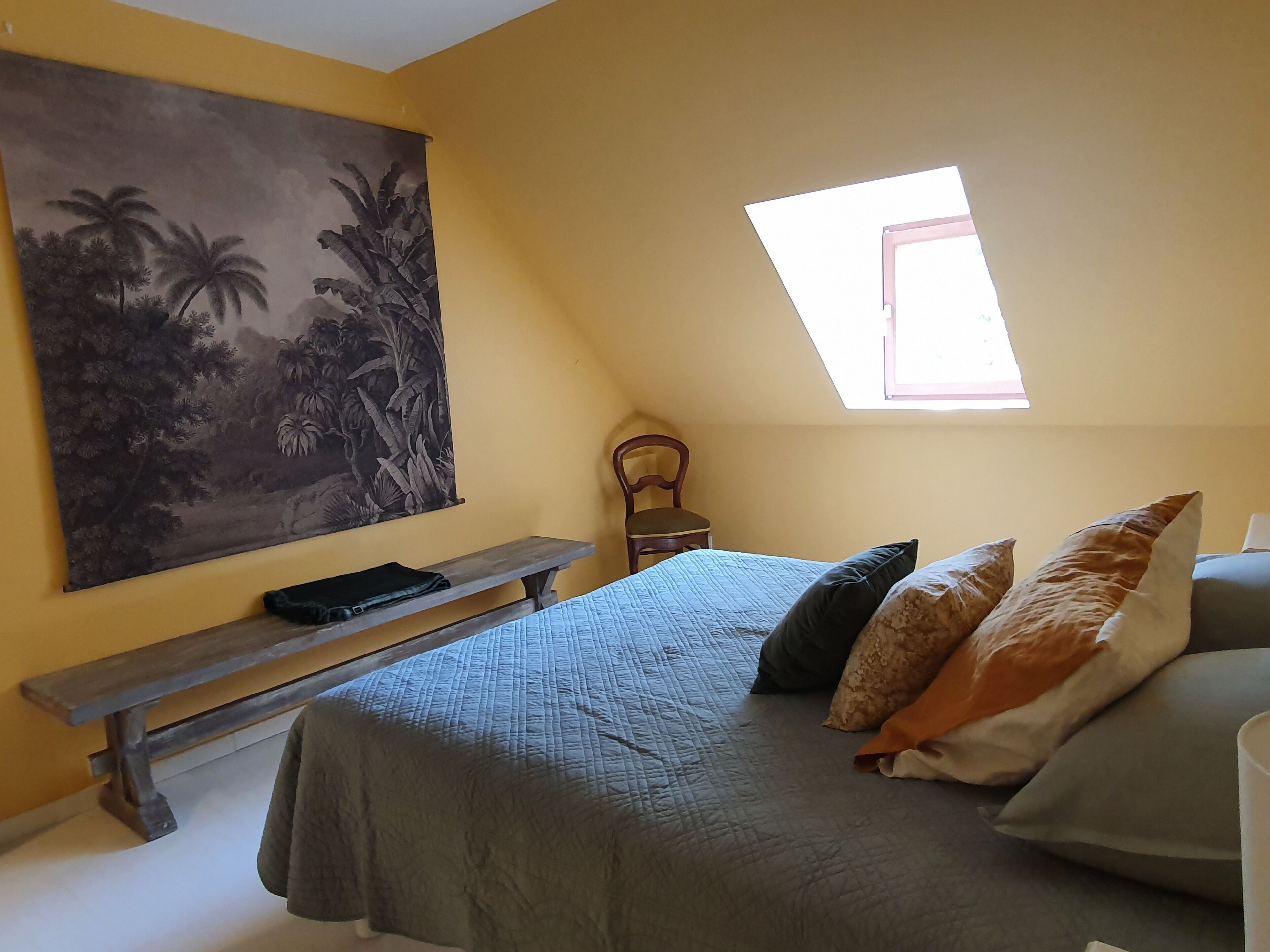 Ein Schlafzimmer mit Bett, Kissen, einer Bank, einem Sessel, einem Gemälde und einem Oberlicht.