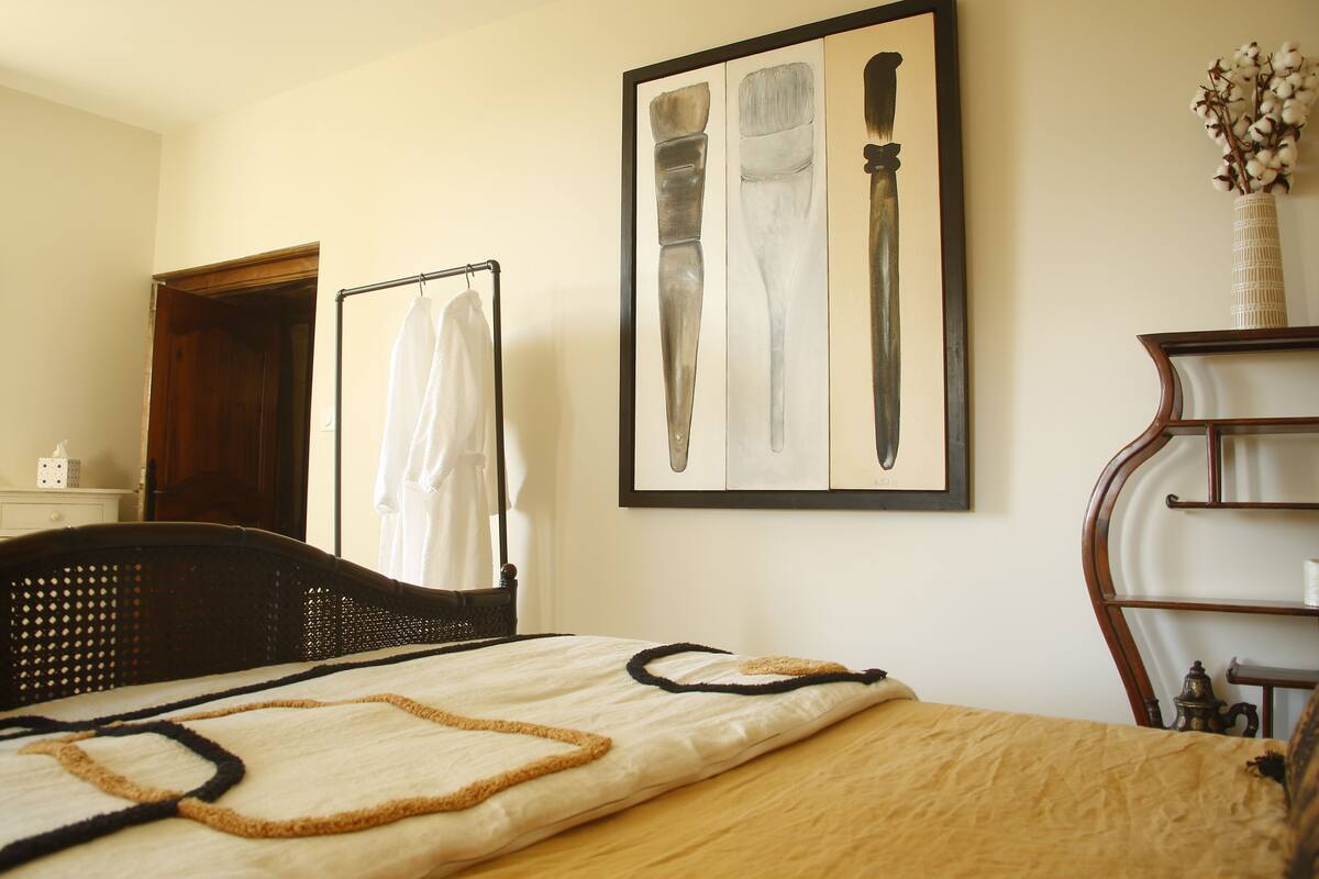 A neatly made bed with a beige and brown bedspread, a framed picture of kitchen utensils on the wall, and a wooden nightstand with a lamp.