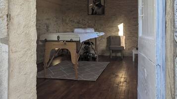 Een rustieke kamer met stenen muren en een houten plafond, een massagetafel, een kleine stoel en een ingelijst schilderij aan de muur.