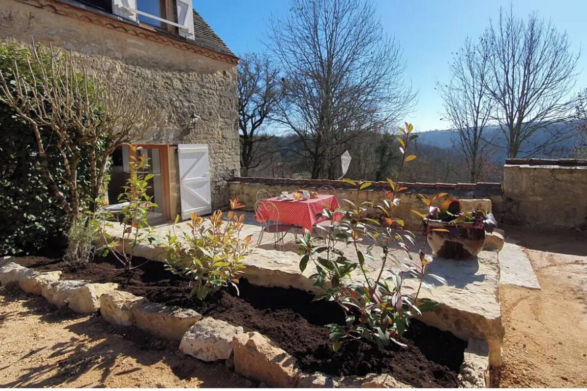 A stone building with a red table and chairs, surrounded by plants and a clear sky.