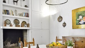 Uma área de refeições com lareira, cadeiras de madeira, uma mesa com uma tigela de frutas e uma estante com livros e objetos decorativos.