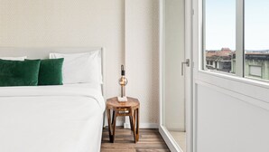 A bedroom with a bed, green pillows, a nightstand with a lamp, patterned wallpaper, and a view of buildings through a window. - Urban Retreat w/ Balcony | Bright & Central Porto (Porto)