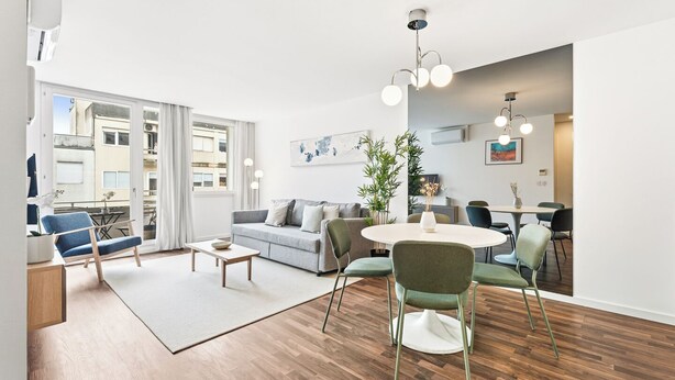 A modern living room with a sofa, coffee table, and dining area with chairs. There is a large window with curtains, a plant, and artwork on the walls. - Urban Retreat w/ Balcony | Bright & Central Porto (Porto)
