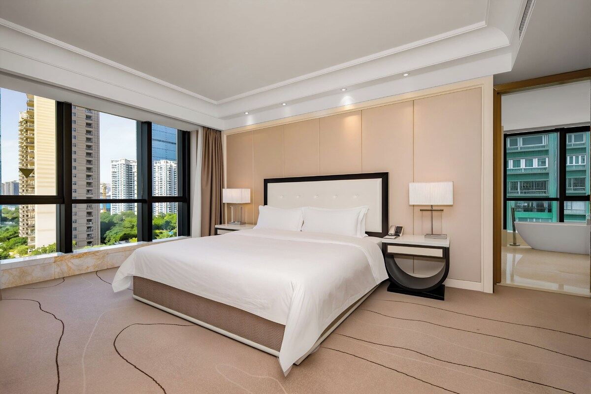 Une chambre d’hôtel moderne avec un grand lit, une table de chevet, une lampe et une vue sur le paysage urbain.