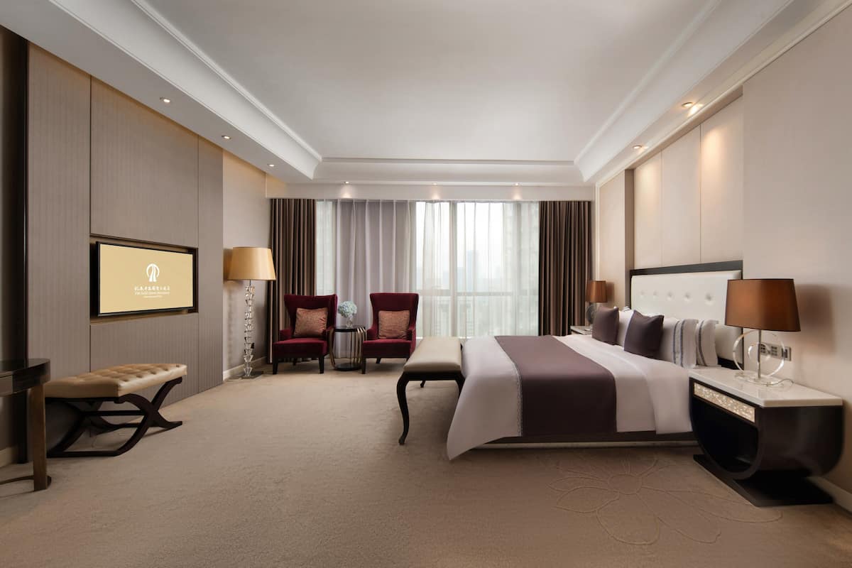 Habitación de hotel moderna con una cama grande, dos sillones, un banco, un televisor y una mesita de noche con lámparas.
