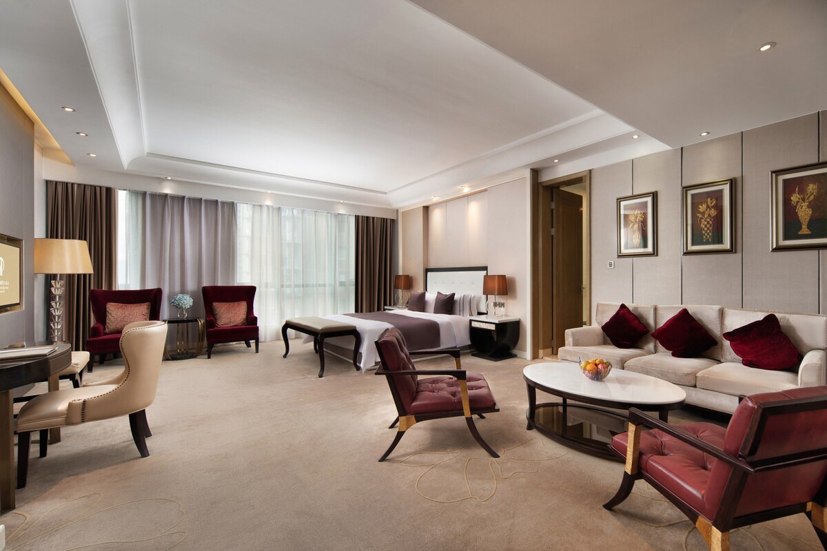 Une chambre d’hôtel spacieuse avec un grand lit, deux fauteuils rouges, un canapé blanc, une petite table ronde avec des fruits et un bureau avec une lampe.