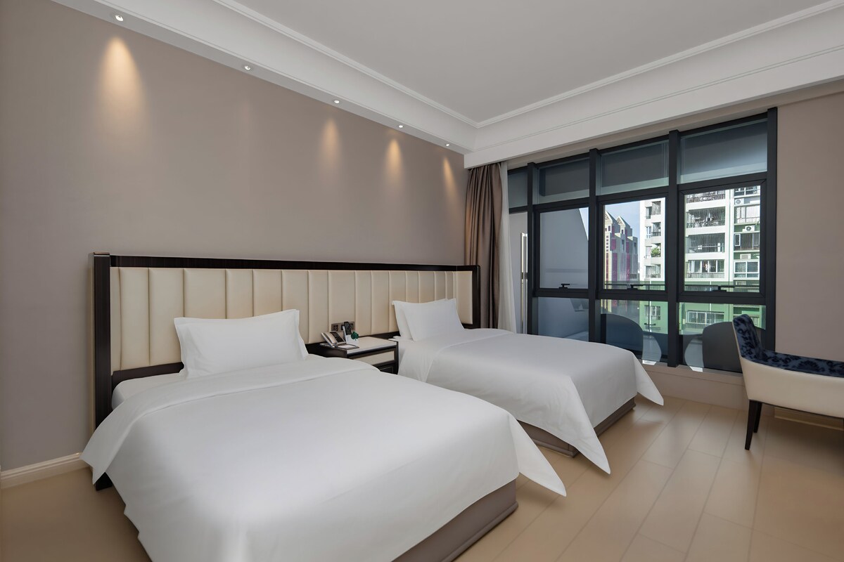 Une chambre d’hôtel avec deux lits, une table de chevet, une chaise et une grande fenêtre donnant sur des immeubles.