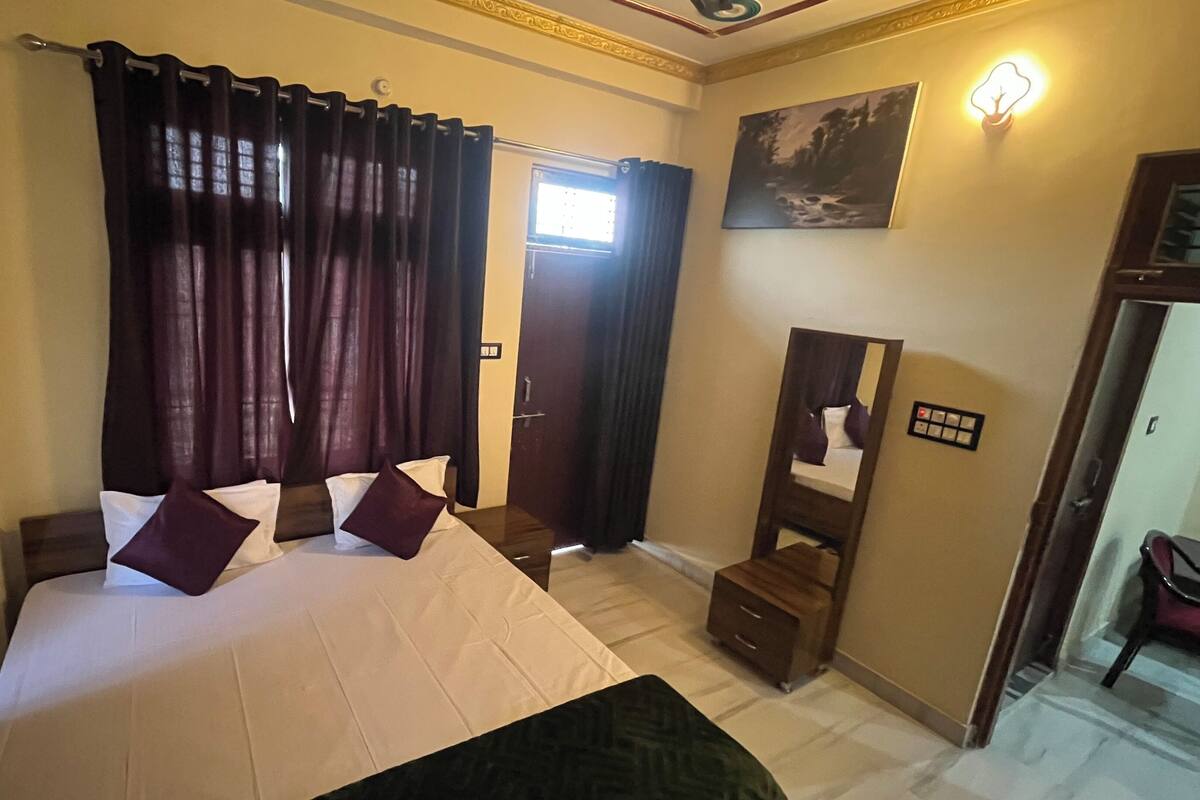 A hotel room with a large bed, dark curtains, a small wooden nightstand, a mirror, and a painting on the wall.