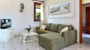 Una sala de estar con un sofá verde, una mesa de centro blanca, un televisor sobre un mueble blanco, una decoración de caballito de mar en la pared y un cuadro enmarcado.