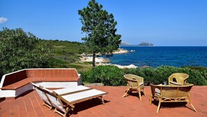Una terraza con una silla lounge blanca, sillas de mimbre y vistas al mar y a la montaña.