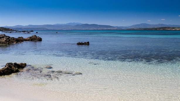 Una playa con agua cristalina, rocas y montañas a lo lejos.