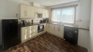 A compact kitchen with light-colored cabinets, a black refrigerator, a built-in oven, a washing machine, and a window with blinds.
