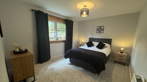 A bedroom with a bed, bedside tables, a nightstand, a window with curtains, a ceiling light, and a radiator.