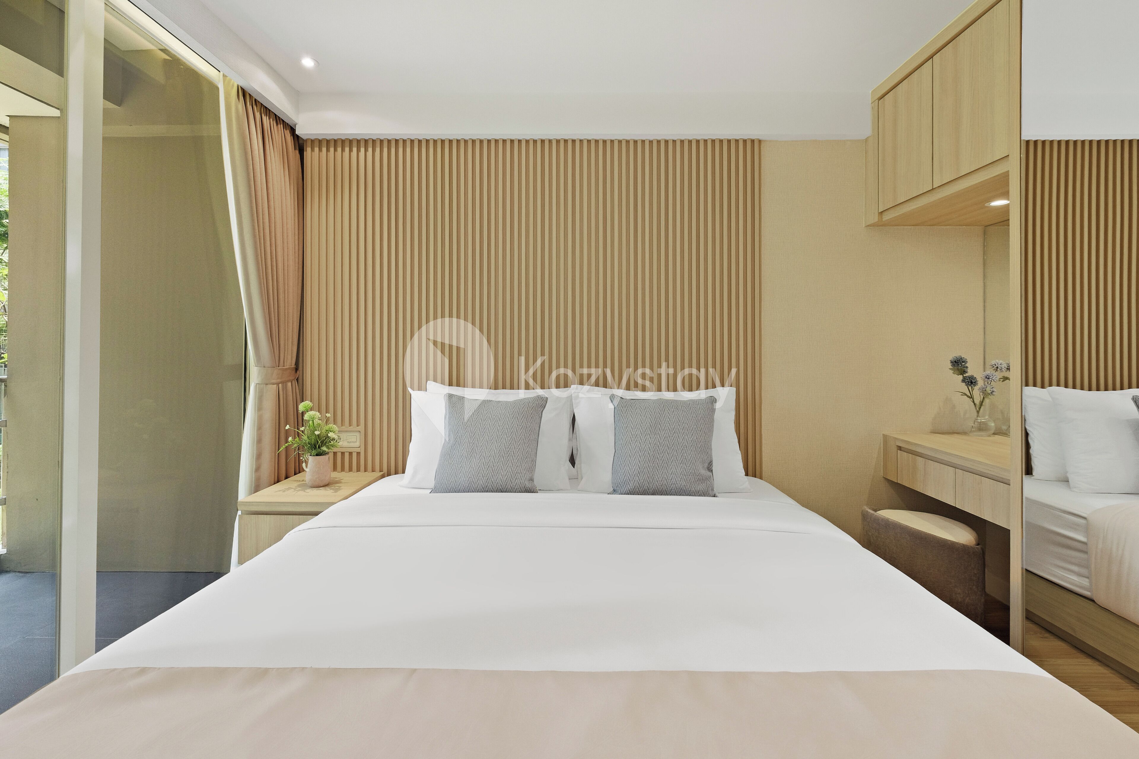 Habitación de hotel moderna con una cama grande, una mesita de noche con lámpara, otra mesita más pequeña con un florero y un armario de madera.