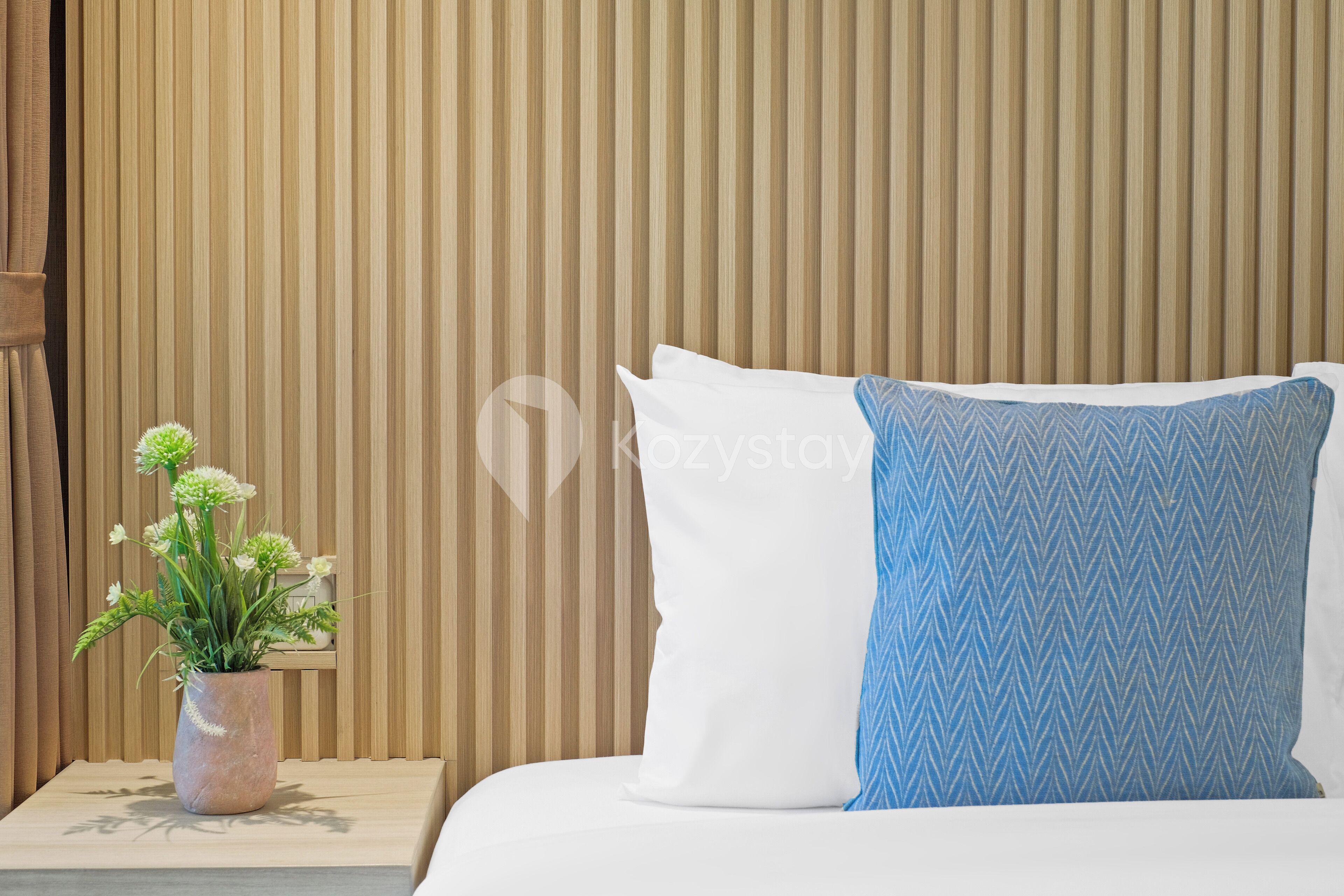 Una cama con almohadas blancas y azules, una pequeña maceta con flores blancas y un muro de listones de madera.