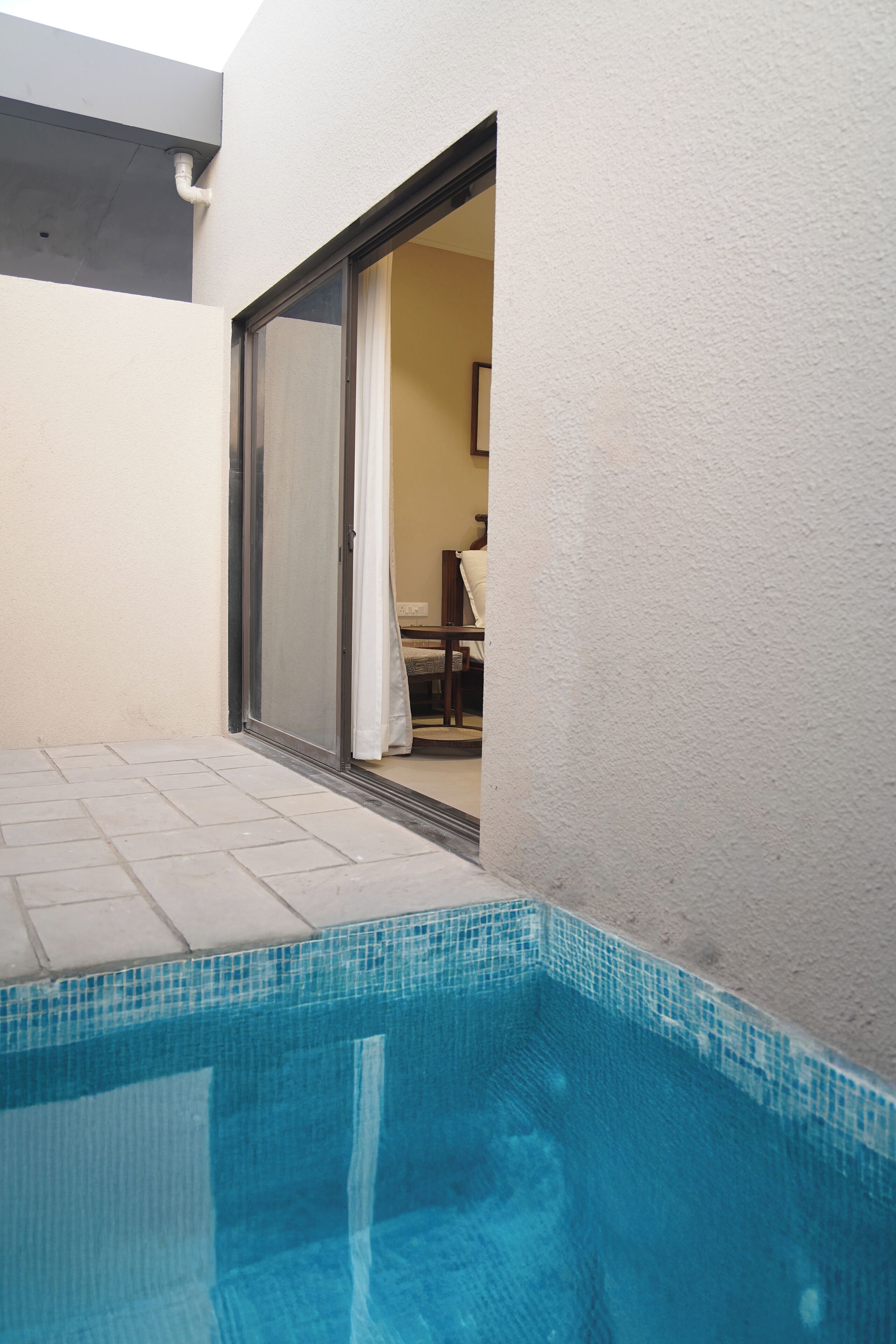 Une piscine avec une terrasse carrelée, une porte-fenêtre coulissante donnant sur une pièce intérieure et un mur extérieur blanc.