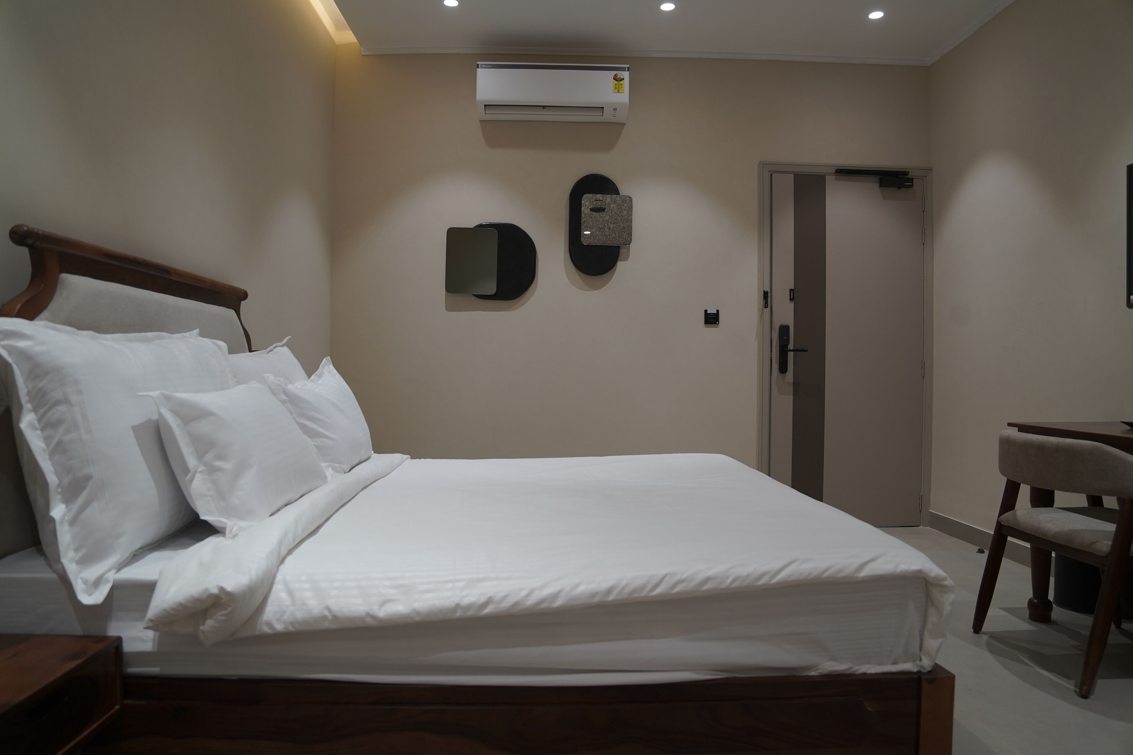 Habitación de hotel con una cama de madera, ropa de cama blanca, un aire acondicionado de pared, un escritorio pequeño con una silla y una puerta.