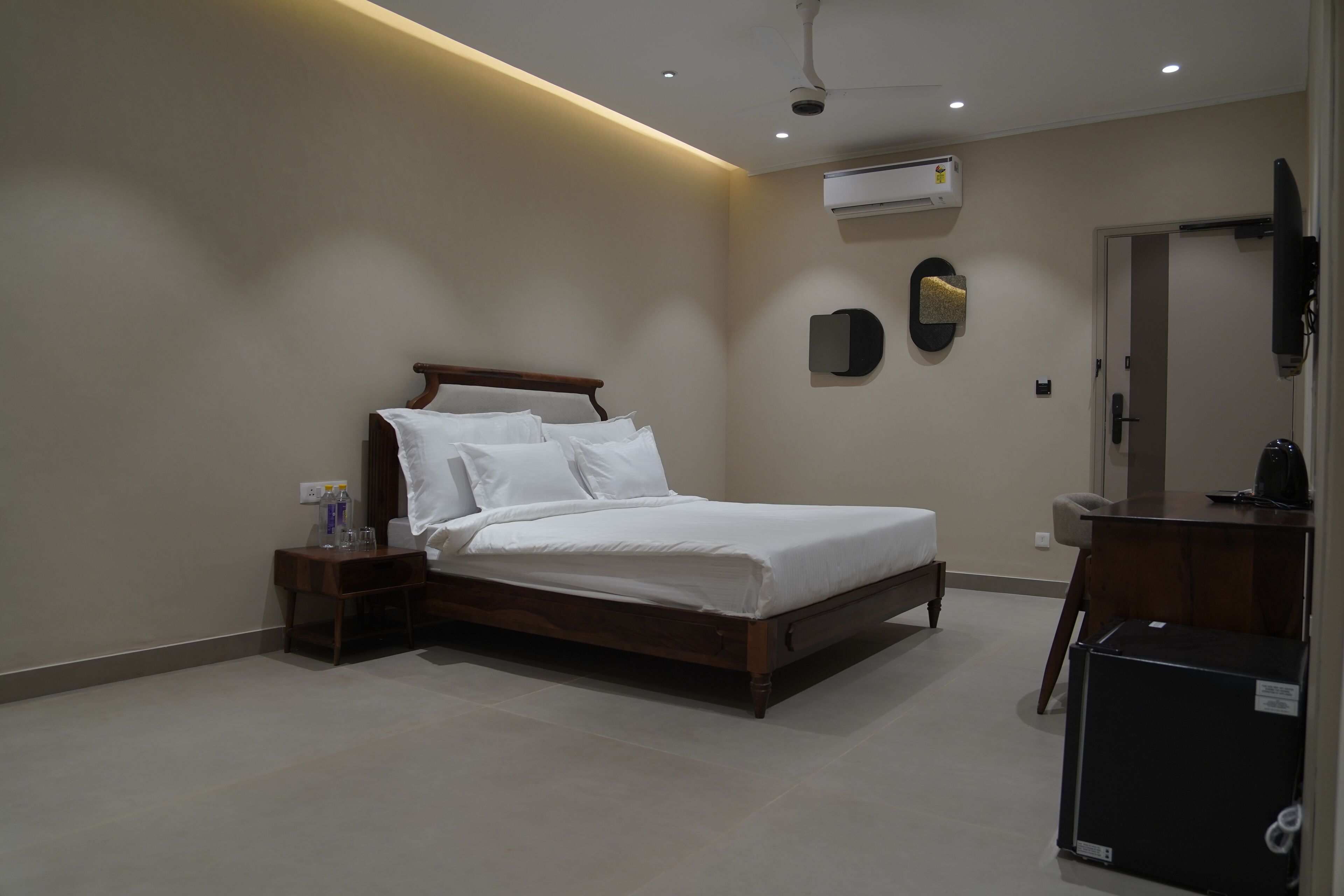 A modern hotel room with a wooden bed, a bedside table with a lamp, an air conditioning unit, and a desk with a chair.