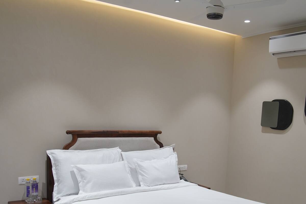 Un lit bien fait dans une chambre d’hôtel, avec une tête de lit en bois, une table de chevet sur laquelle se trouvent une bouteille et un verre, et un ventilateur de plafond.