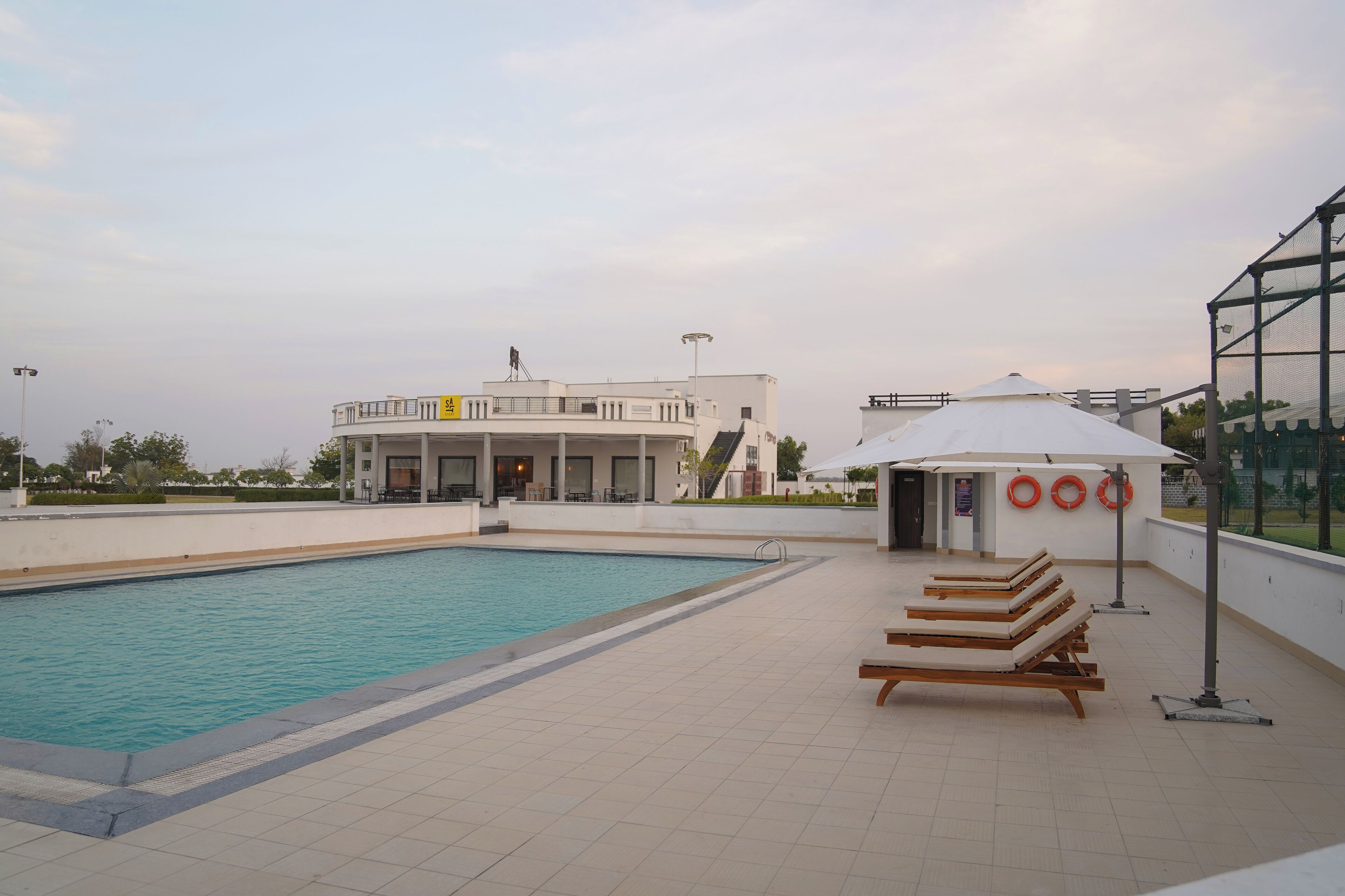 Una piscina con sillones de descanso y sombrillas, un edificio de fachada blanca y boyas salvavidas en la pared.