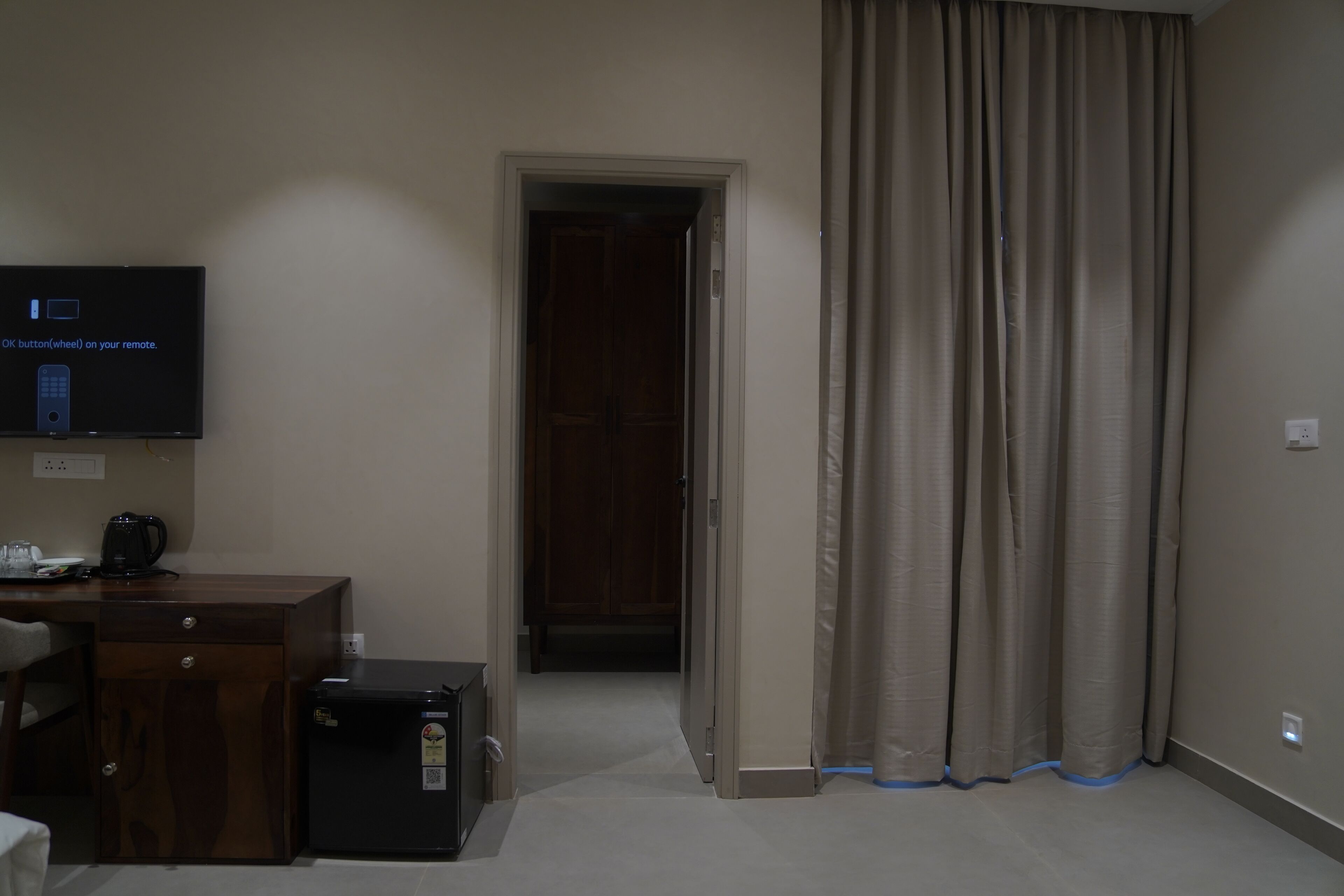 A hotel room with a desk, a television, a wardrobe, a small refrigerator, and a door leading to another room.