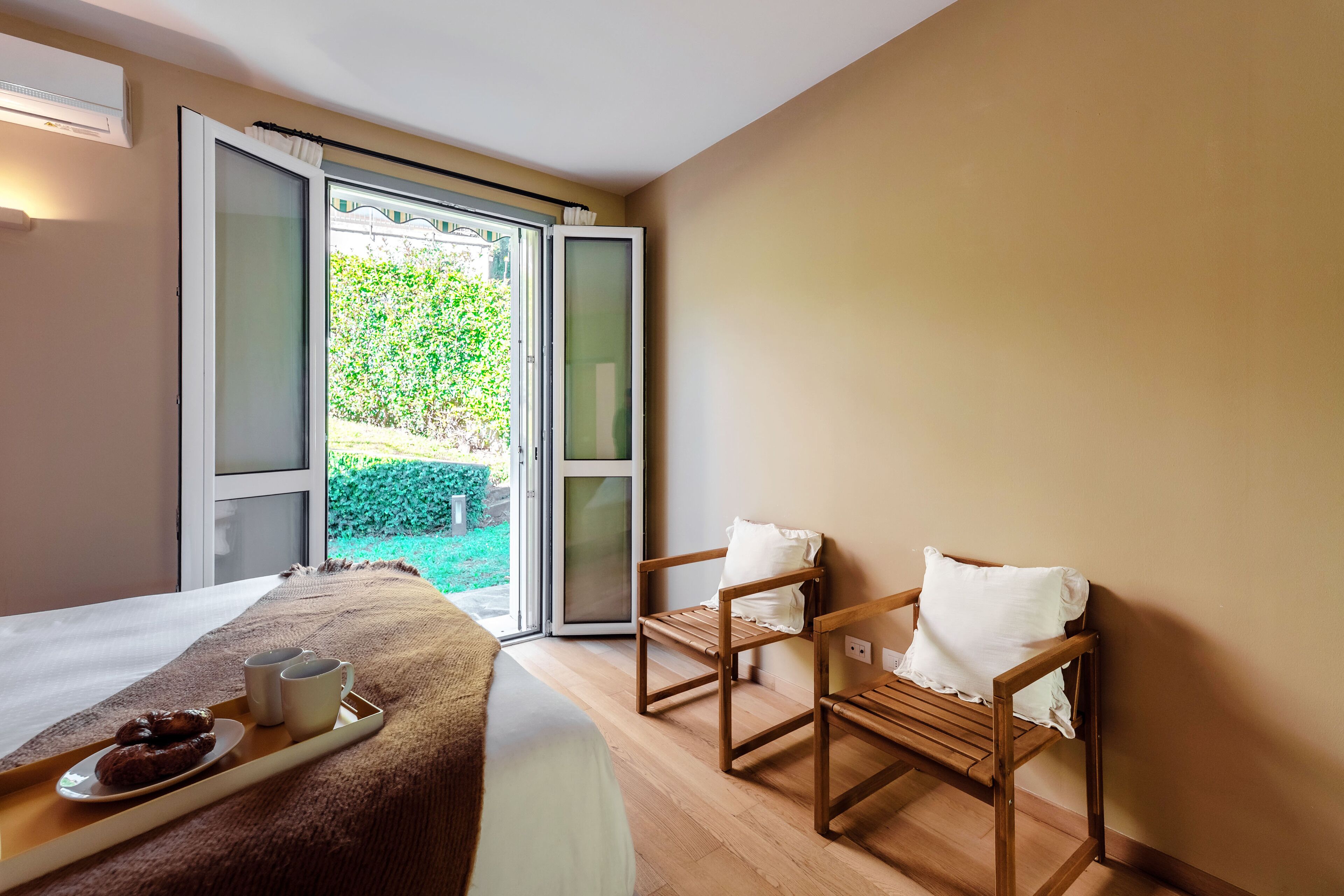 Una habitación con una cama, una bandeja con pasteles y café, dos sillas de madera y un ventanal corredero que da a un jardín.