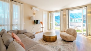A modern living room with a sofa, ottoman, and chair, a flat-screen TV, and a large sliding glass door leading to a balcony with a mountain view. - Residenza di Luce (Griante)