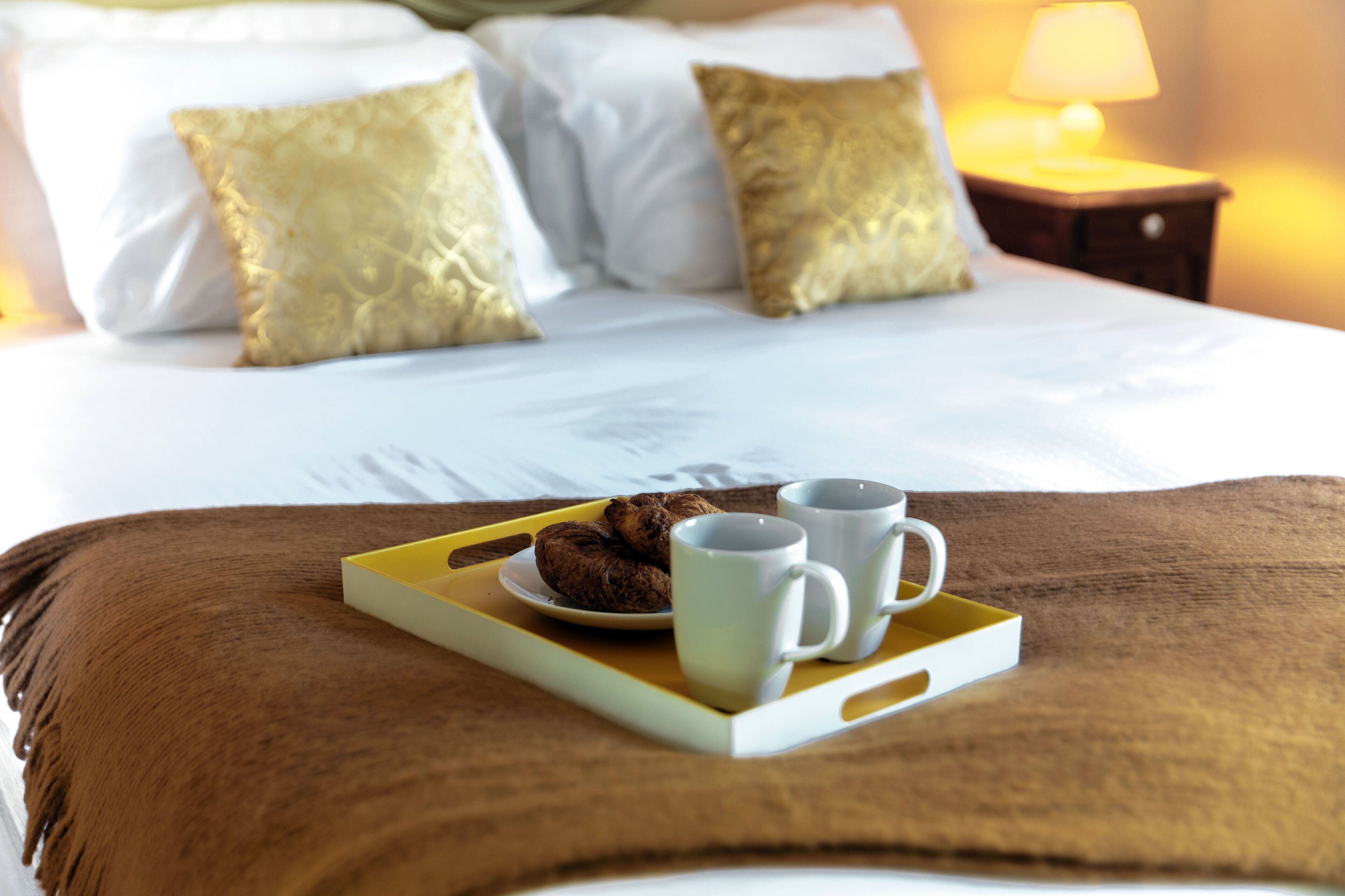 Ein ordentlich bezogenes Bett mit weißen Leinen und goldenen Kissen, ein Tablett mit Keksen und zwei Tassen, sowie eine Lampe auf einem Nachttisch.