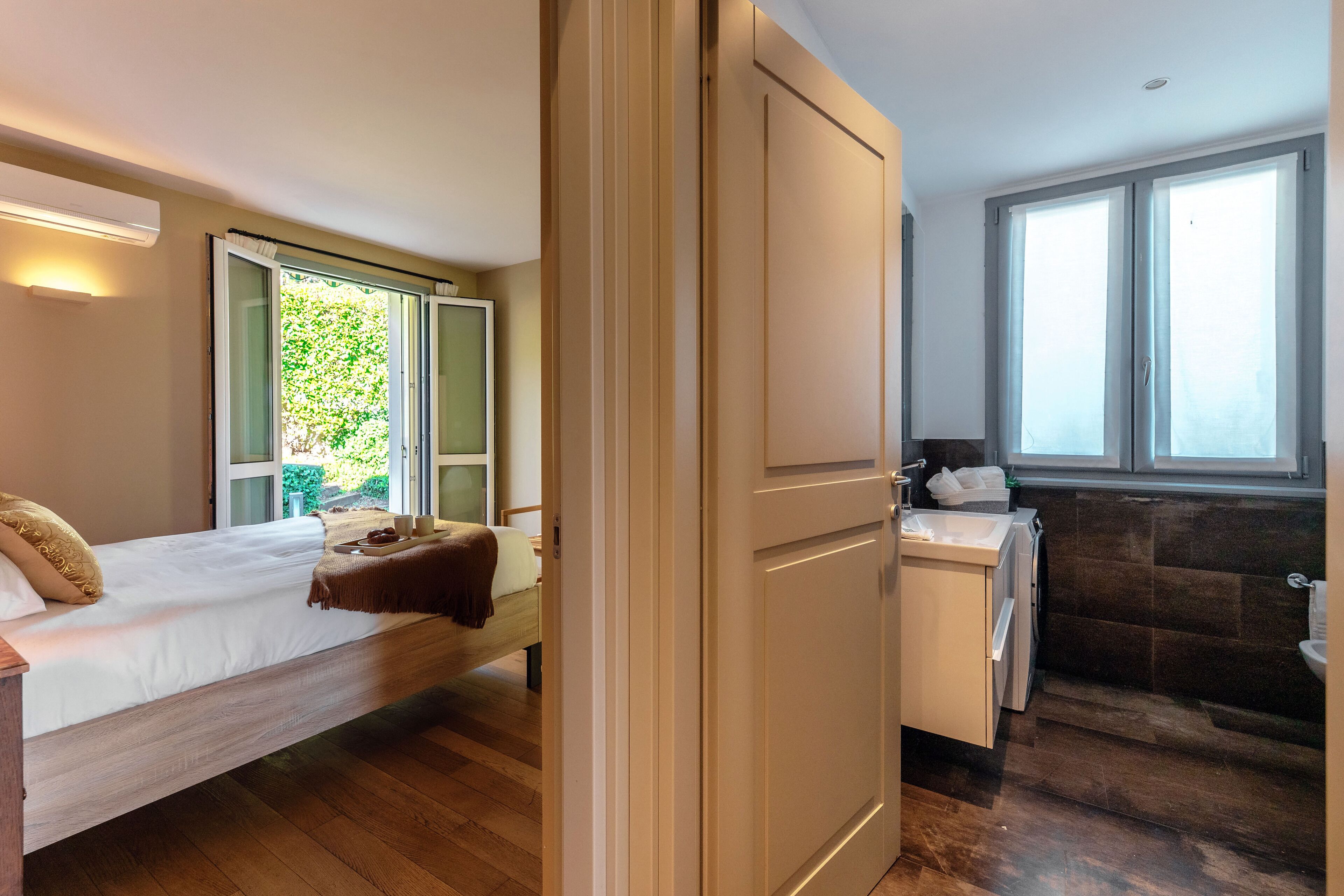 A bedroom with a bed, a bedside table, a window with a view of greenery, and a bathroom with a sink and toilet.