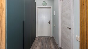 Een smalle gang met een houten vloer, een witte deur met een klok erboven en een lichtschakelaar aan de muur.