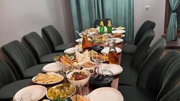 Una mesa de comedor dispuesta con diferentes platos y bebidas, rodeada de sillas negras en una habitación con cortinas color turquesa y una lámpara colgante.
