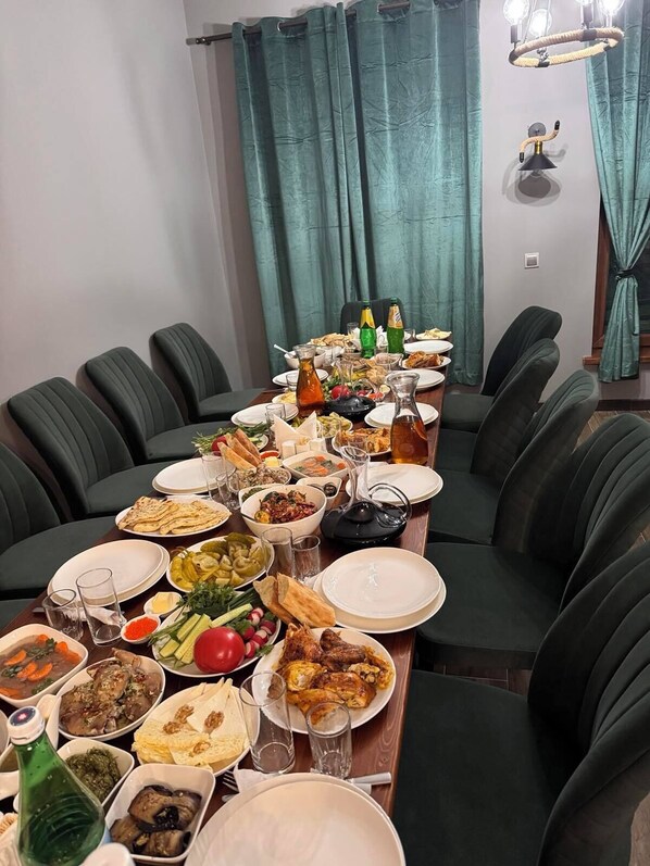Une table à manger dressée avec différents plats et boissons, entourée de chaises noires dans une pièce aux rideaux turquoise et agrémentée d’un luminaire suspendu.