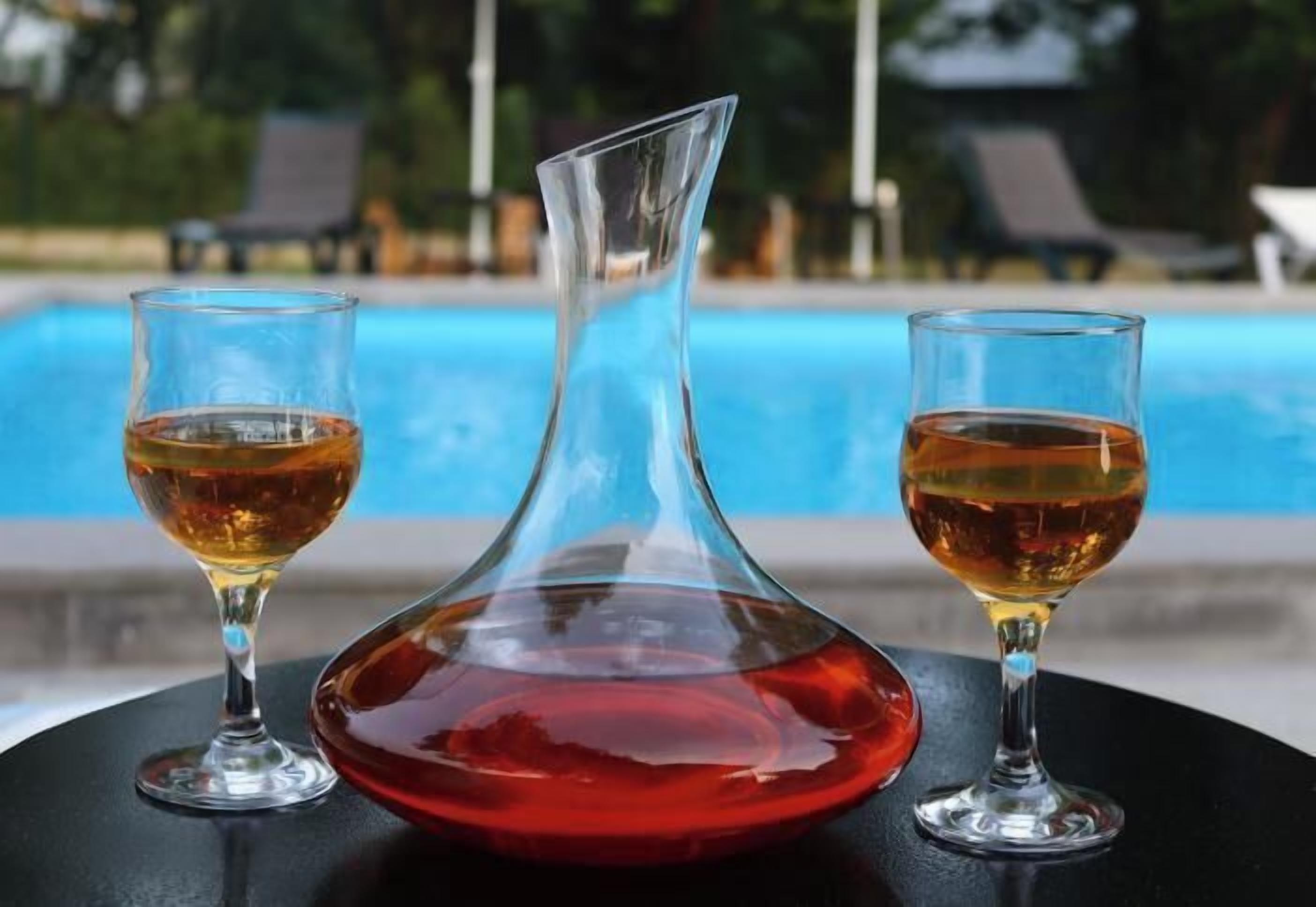 Escena junto a la piscina con una mesa que sostiene tres vasos con líquido color ámbar, probablemente una bebida, y una jarra de vidrio rojo.
