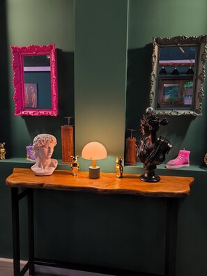Una habitación con una mesa de madera que exhibe un busto, dos figuritas doradas, una lámpara y un espejo con marco rosa.