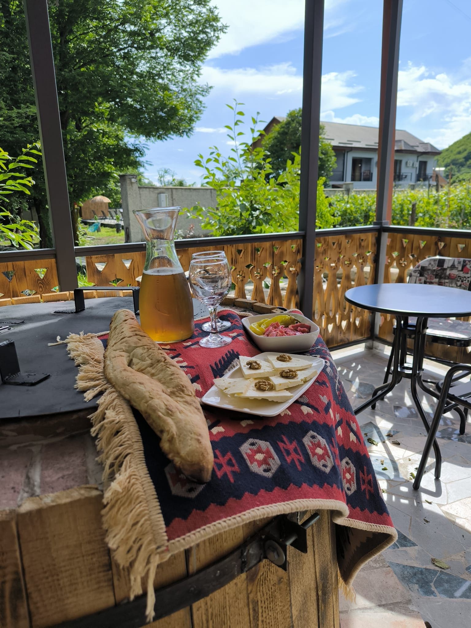 A table with a bottle of drink, a glass, a bowl of food, and a plate with cheese and nuts on a patterned tablecloth.