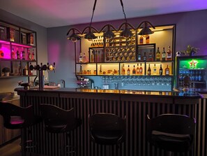 Zona de bar con mostrador, estantes llenos de botellas de licor, vasos colgantes y un letrero de neón.