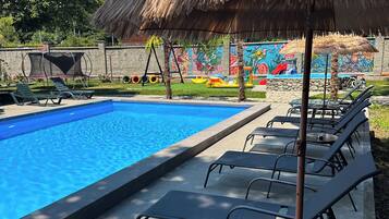 Una piscina con sillas de descanso y una sombrilla de paja, rodeada por un mural y vegetación.