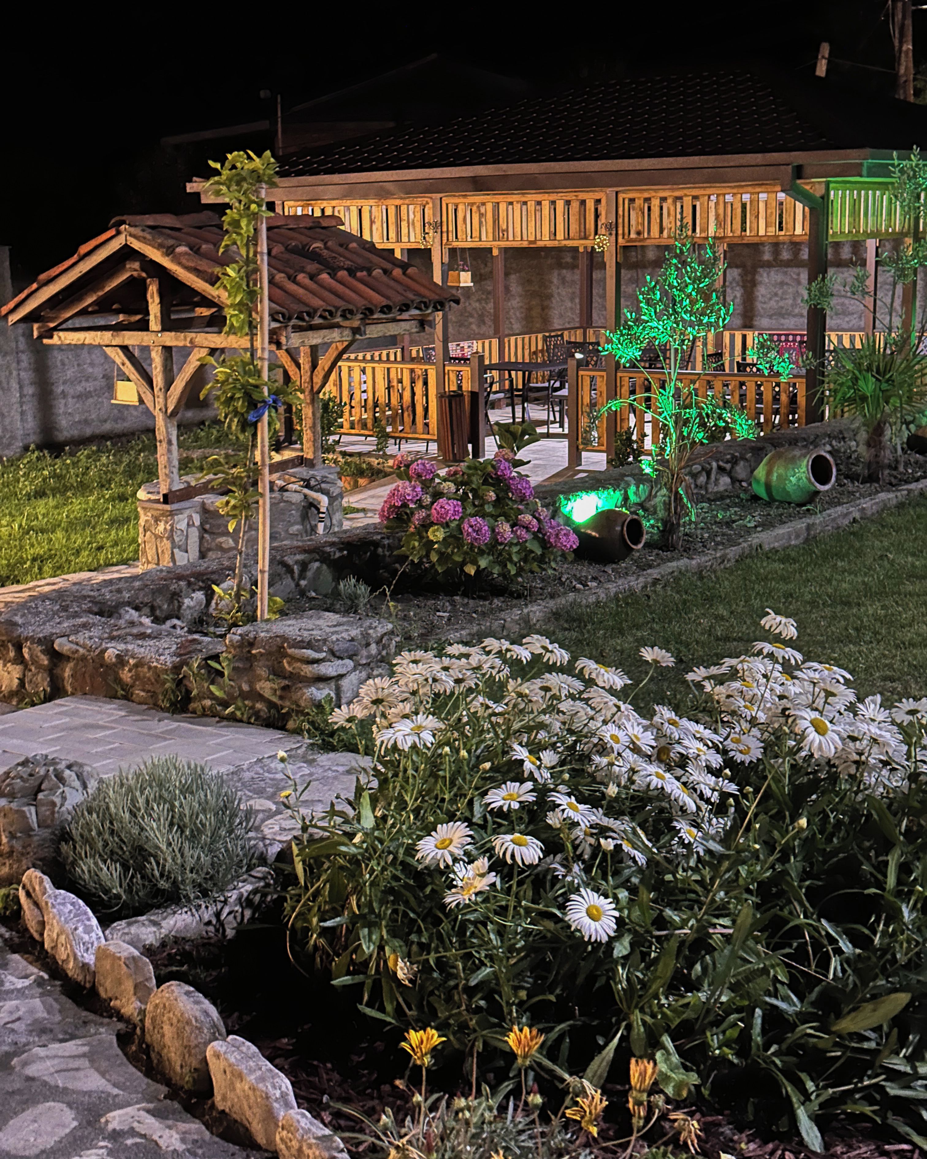 A house with a wooden porch, a stone pathway, and a garden with flowers and a green-lit tree.
