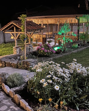 Una casa con porche de madera, un sendero de piedra y un jardín con flores y un árbol iluminado de verde.