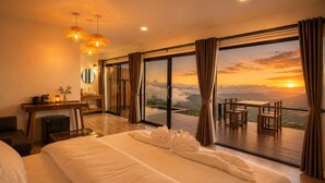 A hotel room with a large bed, a desk, a chair, and a view of the sunset through the window. - Cozy Homey Doichang (Mae Suai)