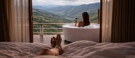 A bedroom with a large bed, a bathtub, and a balcony with a view of mountains.