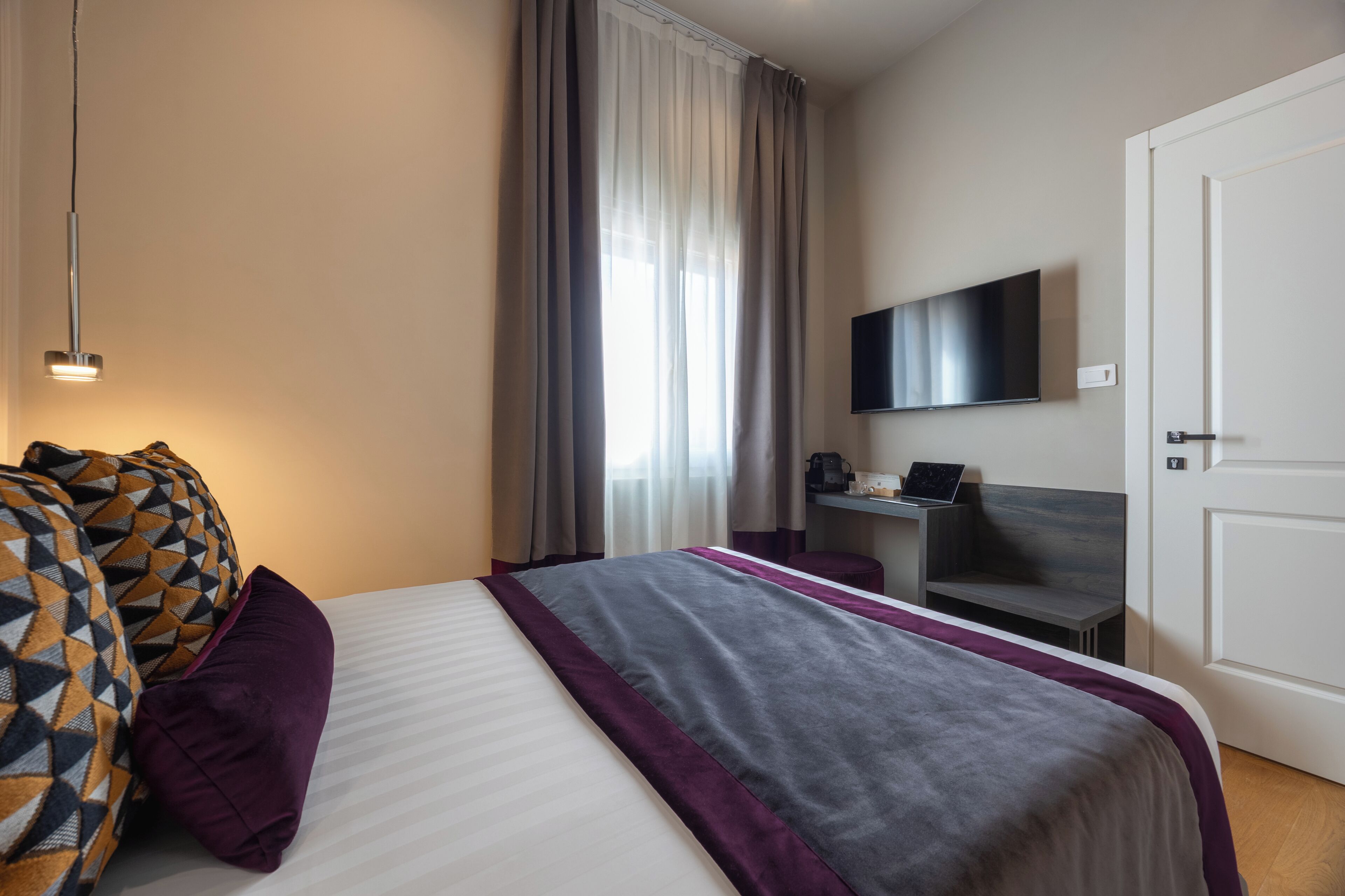 Una habitación de hotel con cama, escritorio con computadora portátil, televisor en la pared y una puerta.